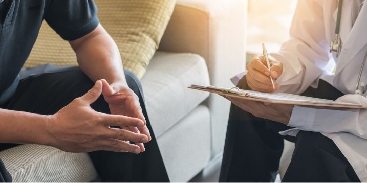 A person sits on a couch talking to a doctor in a white coat who is taking notes on a clipboard, conveying a serious and professional tone.