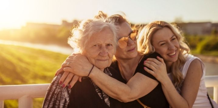 Three women outdoors embrace warmly on a sunny day. An elderly woman and two younger women share smiles, conveying a tone of love and togetherness.