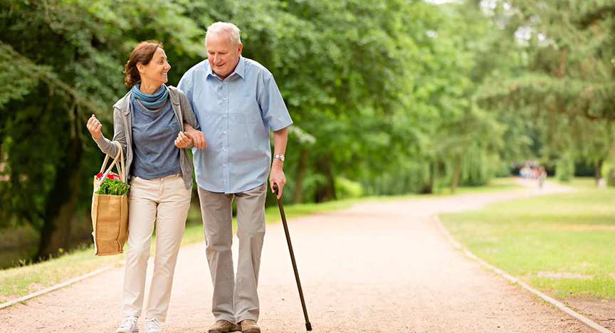 A woman and an elderly man with a cane walk on a park path, smiling. She carries a grocery bag.