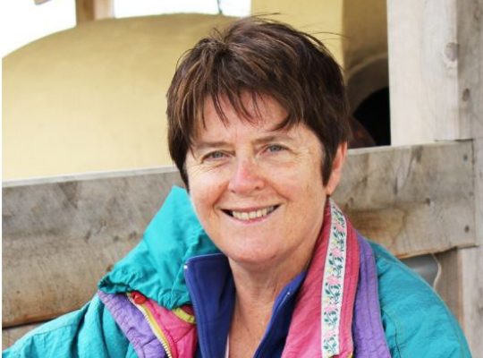 Woman with short brown hair smiling warmly, wearing a colorful jacket. She is outdoors, leaning on a wooden structure.