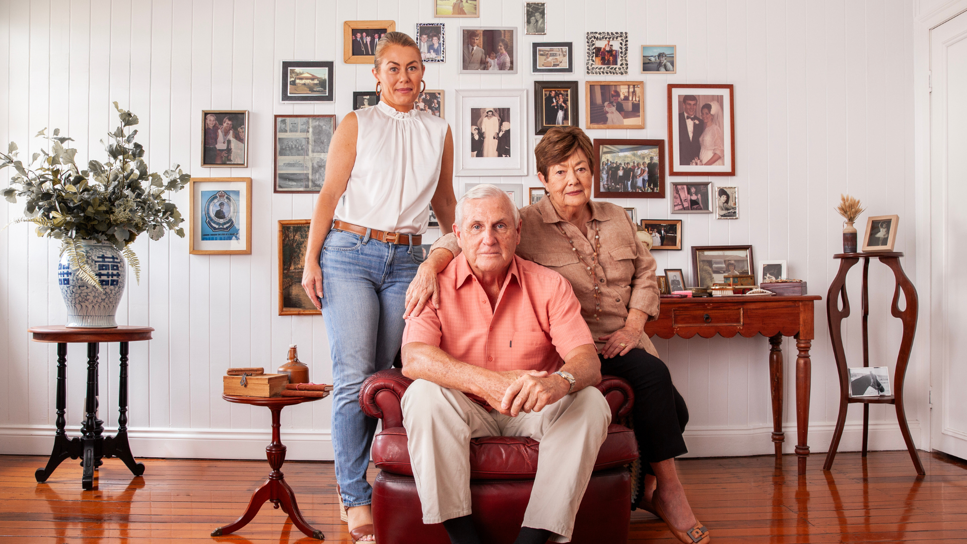 A family of three poses in a cozy room with white walls adorned with numerous framed photos. The older man sits in a red armchair, flanked by the older woman and younger woman standing beside him.