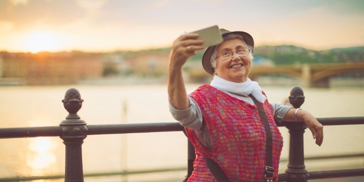 Elderly woman smiling and taking a selfie by a riverside, wearing a hat and a red sweater. The sunset casts a warm glow, creating a joyful atmosphere.